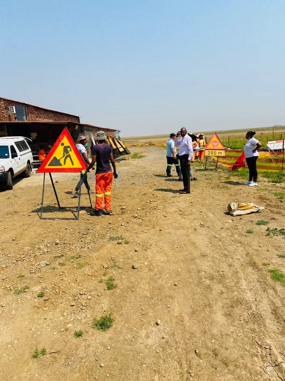 Workers setting up road construction signs in a rural area with a dirt road and vehicles.