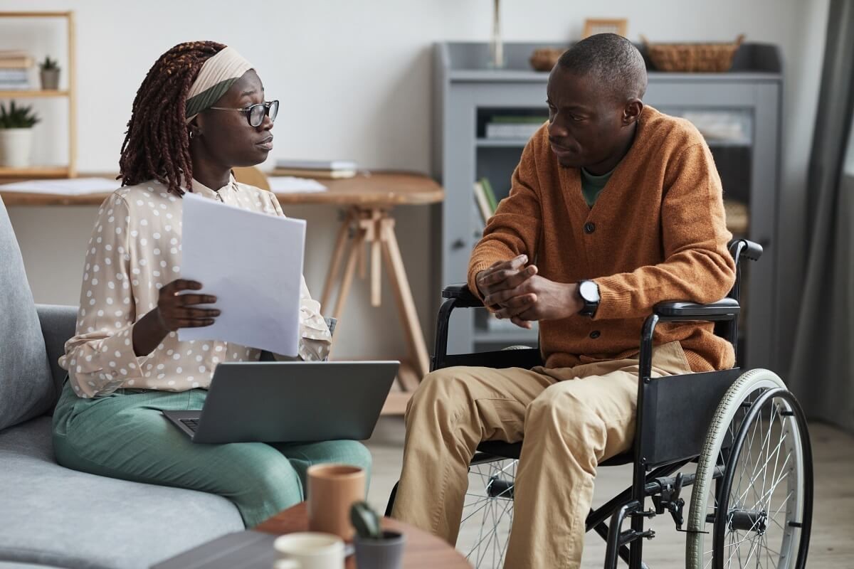 Woman with papers talking to a man in a wheelchair in a cozy living room setting.