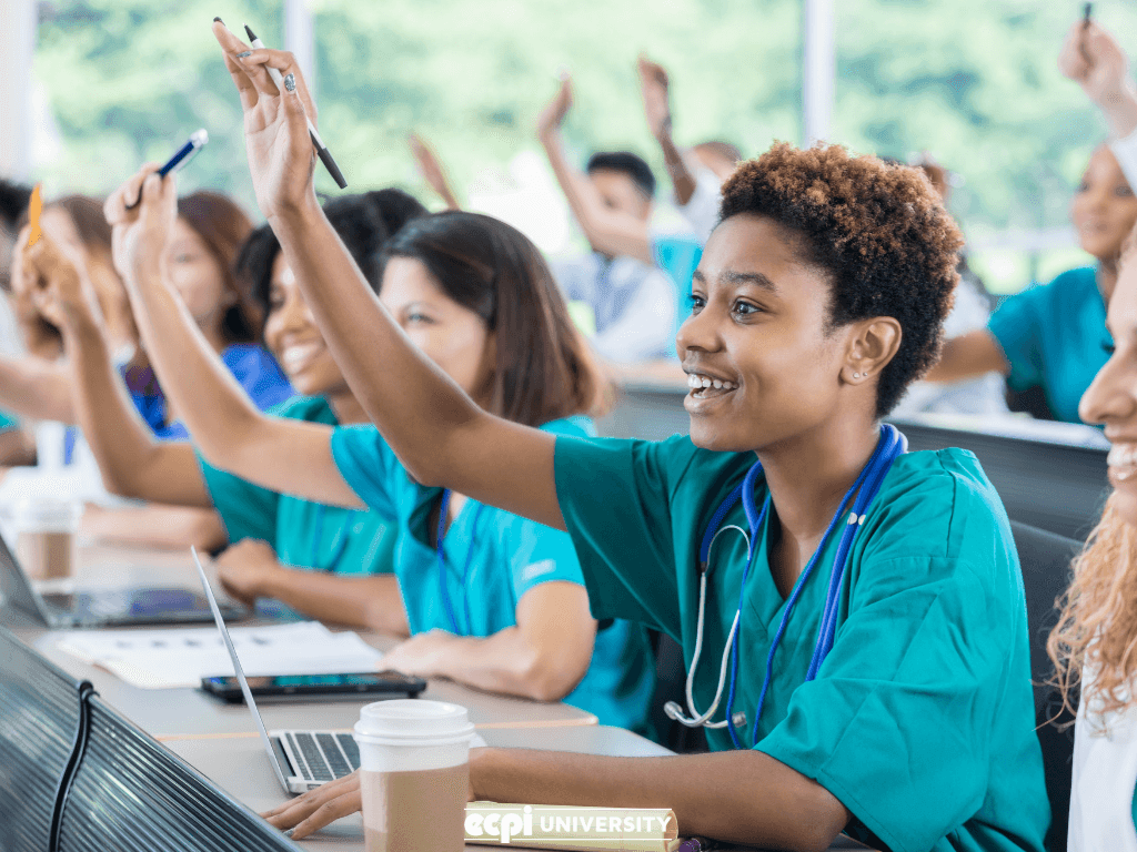 Nursing students in scrubs enthusiastically raising hands during a classroom lecture.