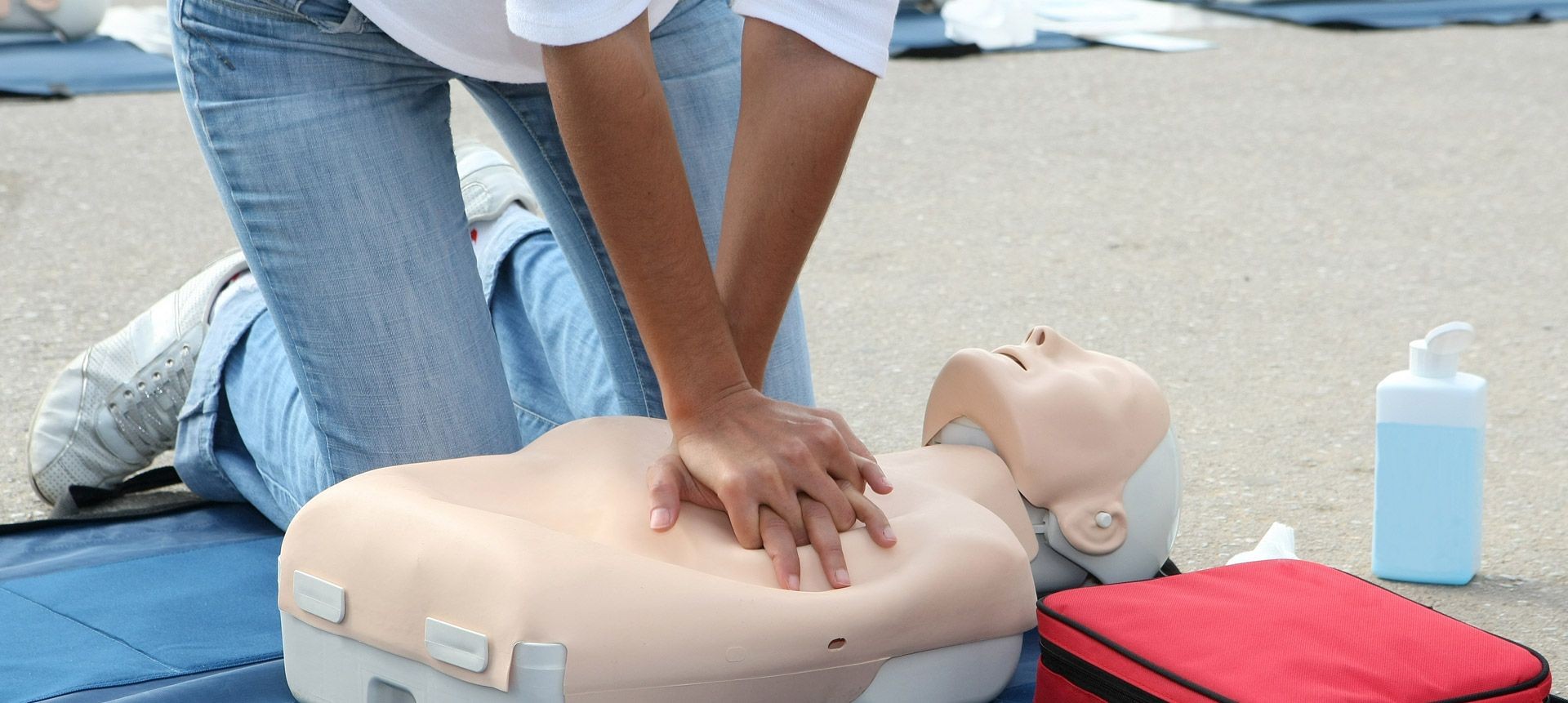 Person practicing CPR on a training mannequin outdoors, with a red bag and sanitizer bottle nearby.